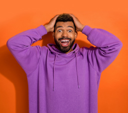 Die Abbildung zeigt einen strahlenden, begeisterten jungen Mann, der in einem lila Sweatshirt vor orangenem Hintergrund steht und beide Händen an den Kopf hält